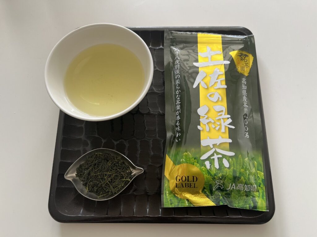 黒い角盆の上に、淡い黄緑色の煎茶を注いだ白い湯呑、茶葉をのせた茶さじ、JA高知県「土佐の緑茶」の袋を並べたところ